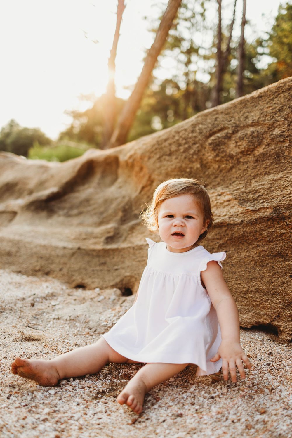 A little one in white dress sits against sunlit coastal rocks.