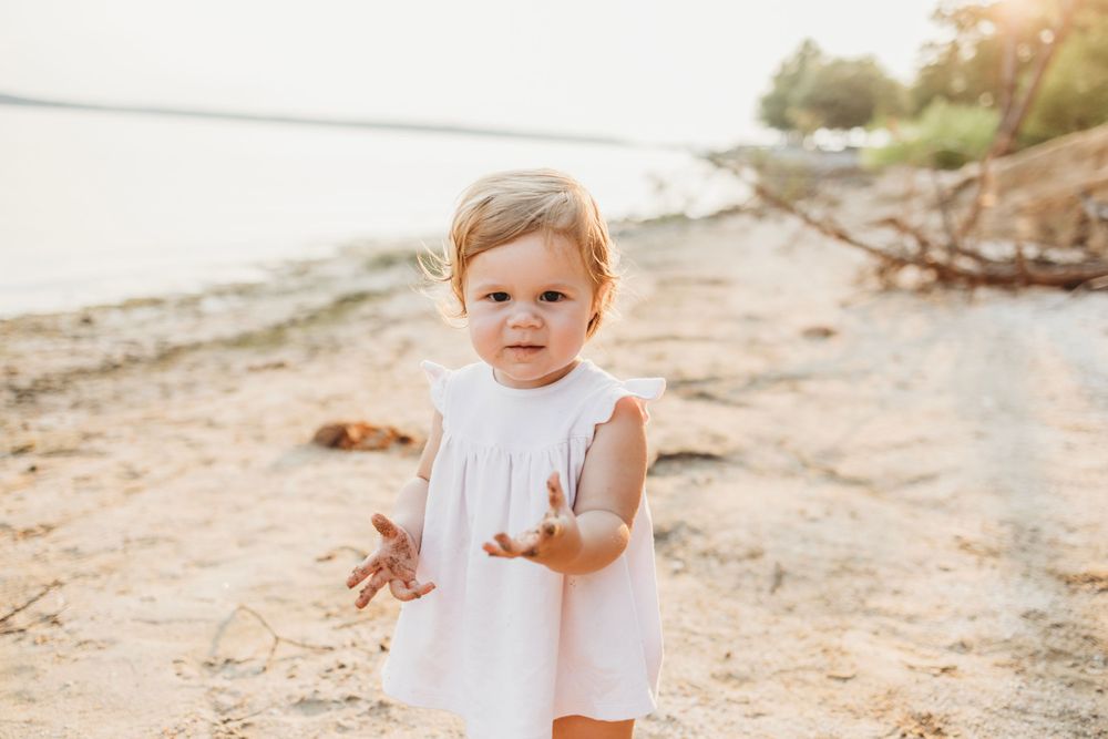 A small child in white dress stands on beach at sunset.