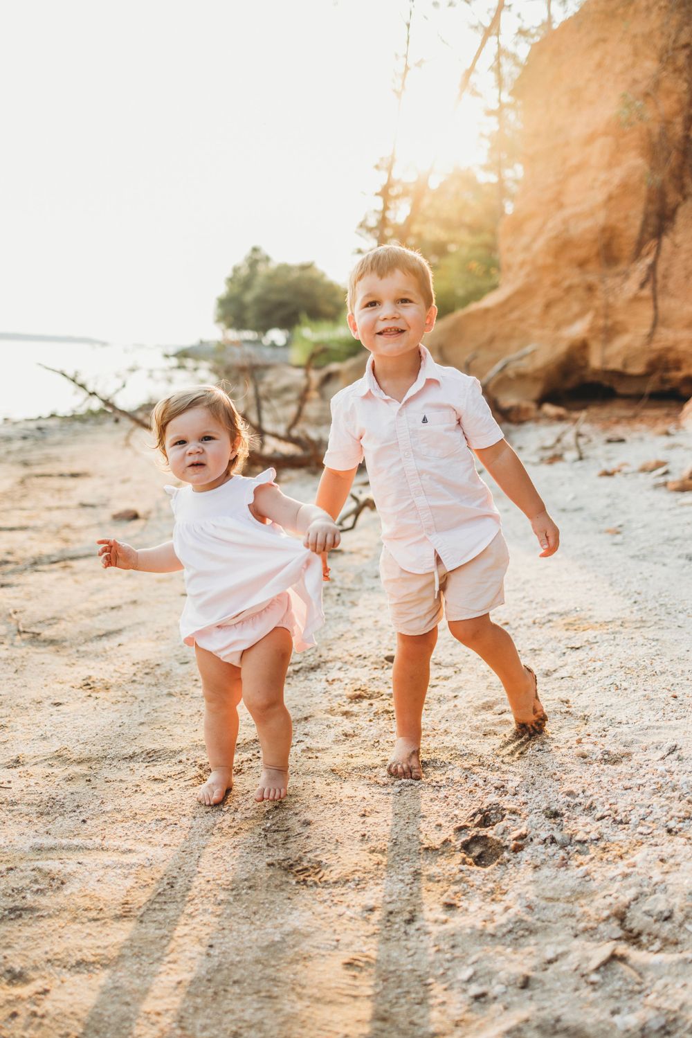 Two children in white outfits run joyfully along a sandy beach with cliffs behind them.