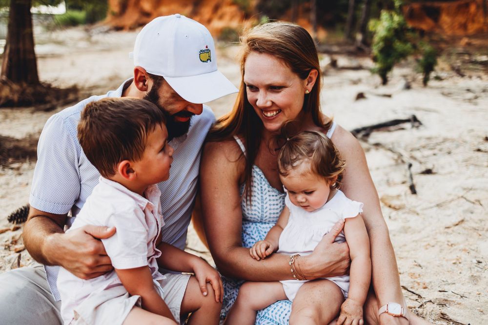 A family shares a tender moment together sitting on a sandy beach near orange cliffs.