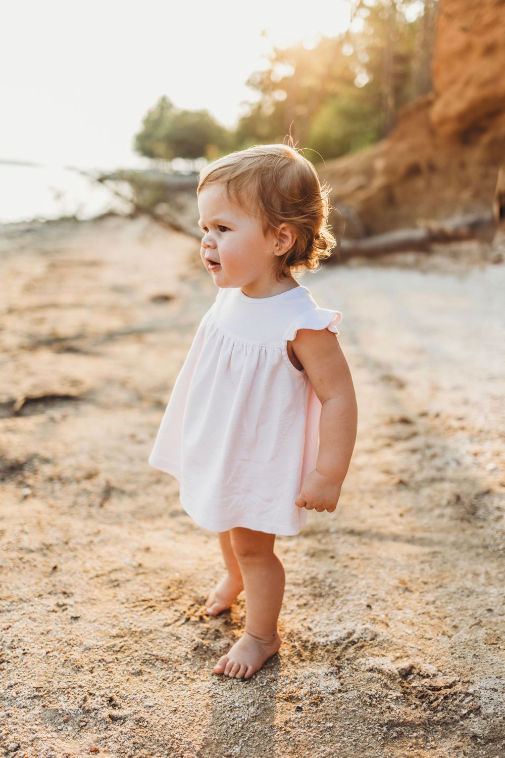 A small child in a white dress stands barefoot on a beach with orange cliffs in the background.