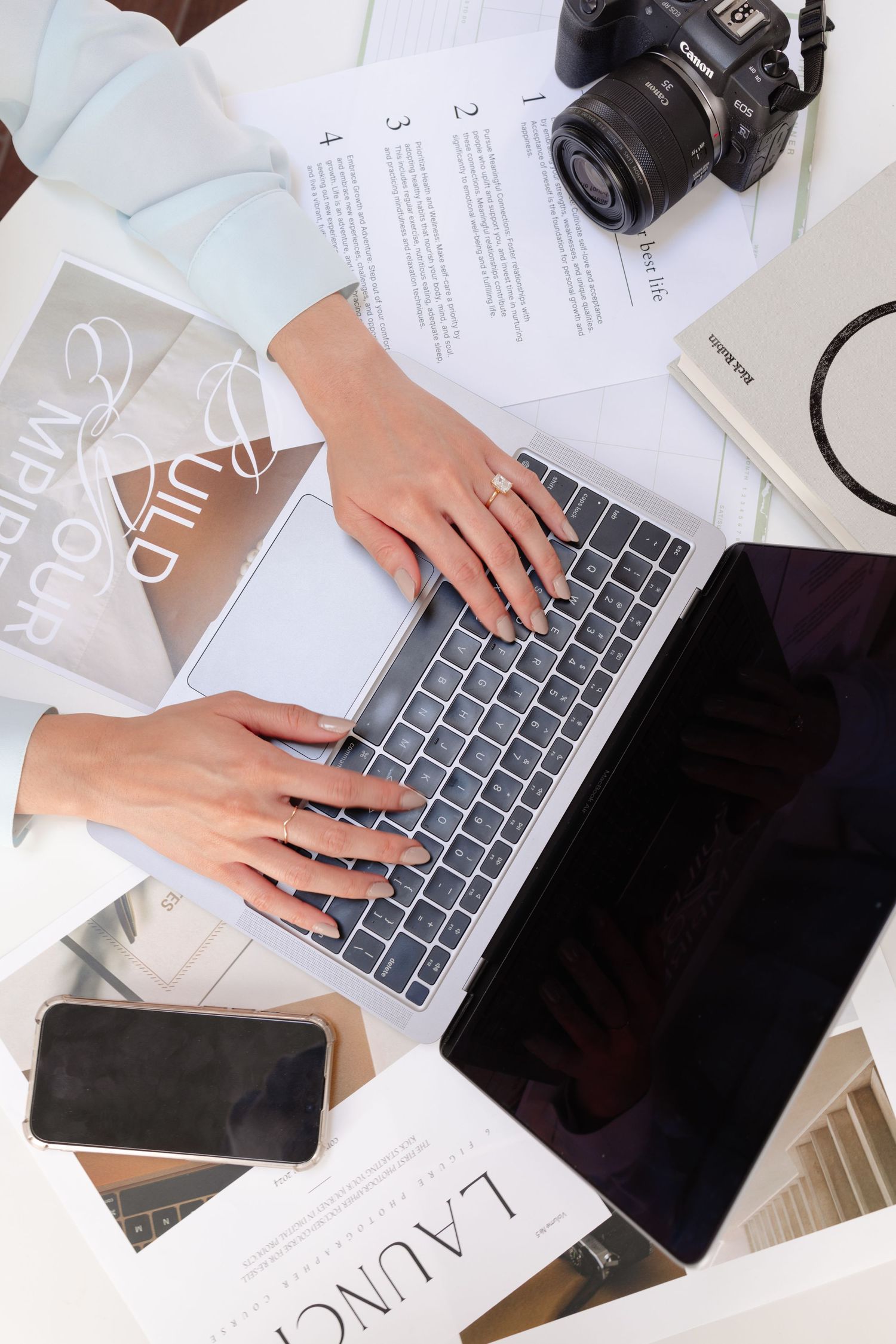 Overhead view of hands typing on laptop with camera and business materials on white desk.