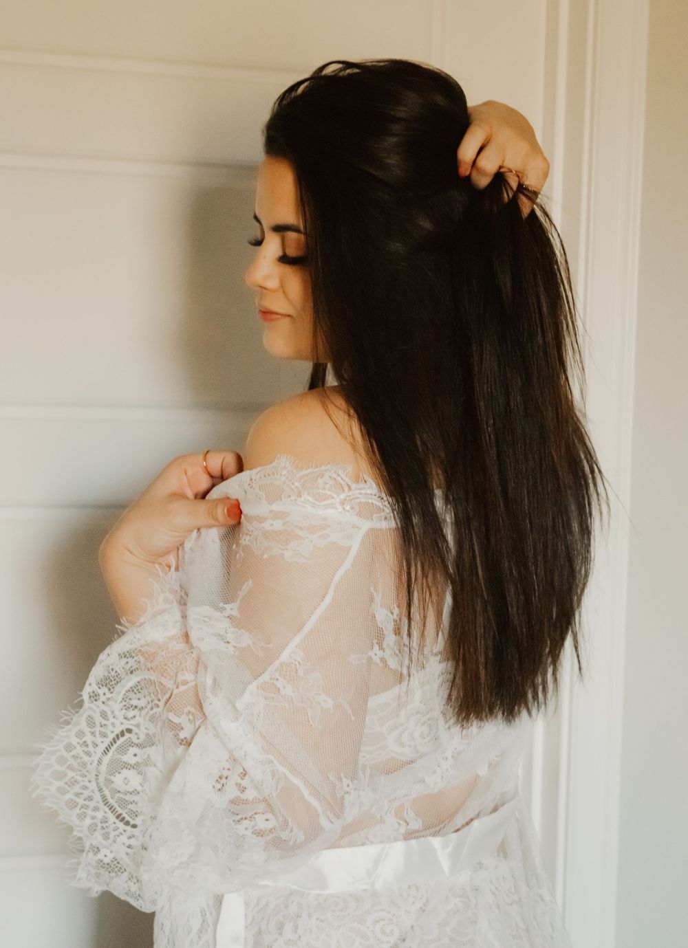 Woman in white lace robe runs fingers through long dark hair against white wall in soft natural light.