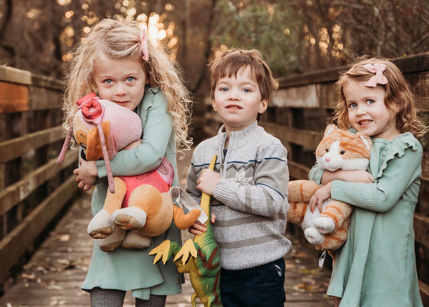 Three children stand together on a wooden bridge holding stuffed animal toys.
