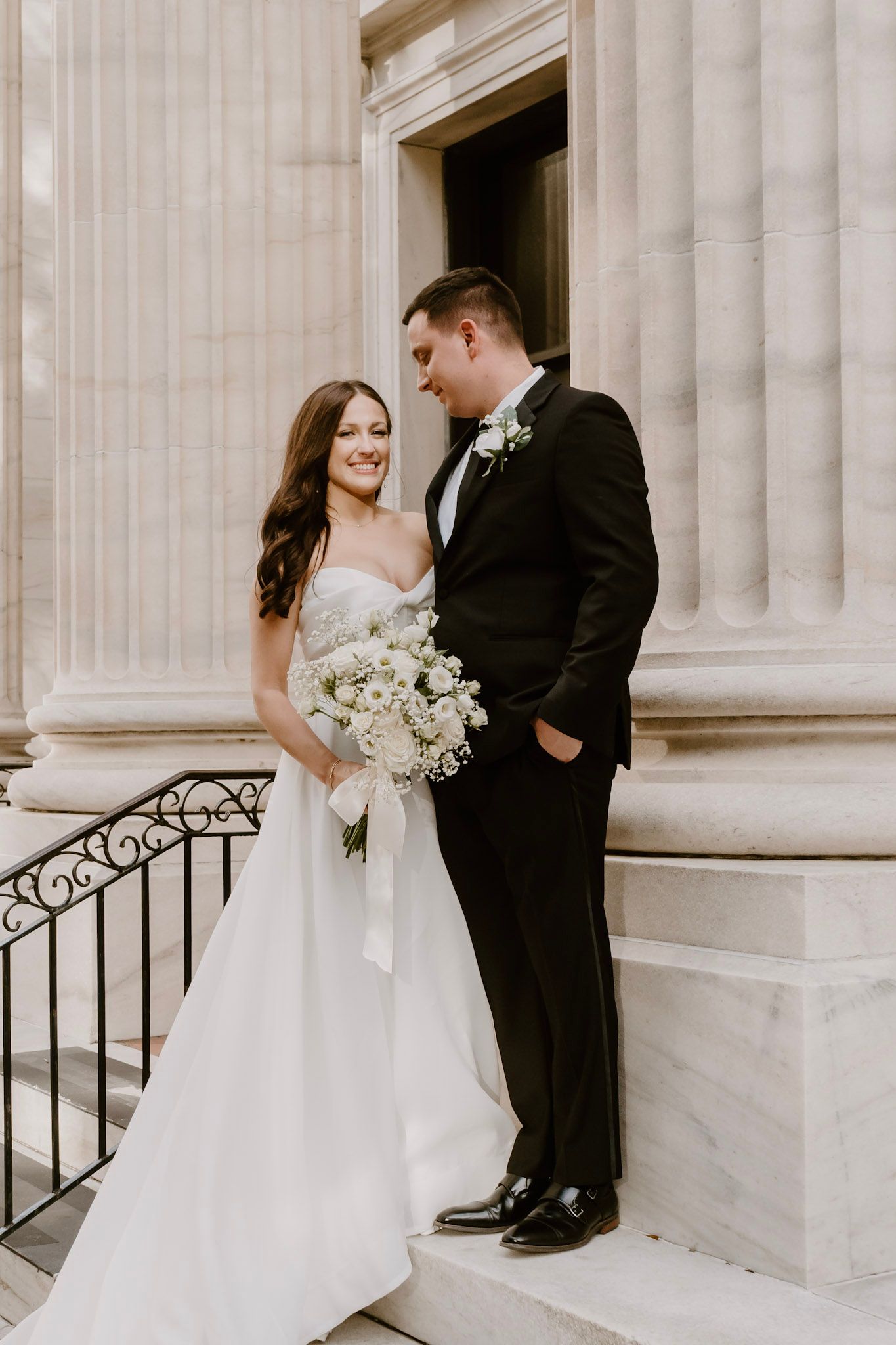 A bride and groom share an intimate moment on marble steps with classic columns, she holds a white floral bouquet.