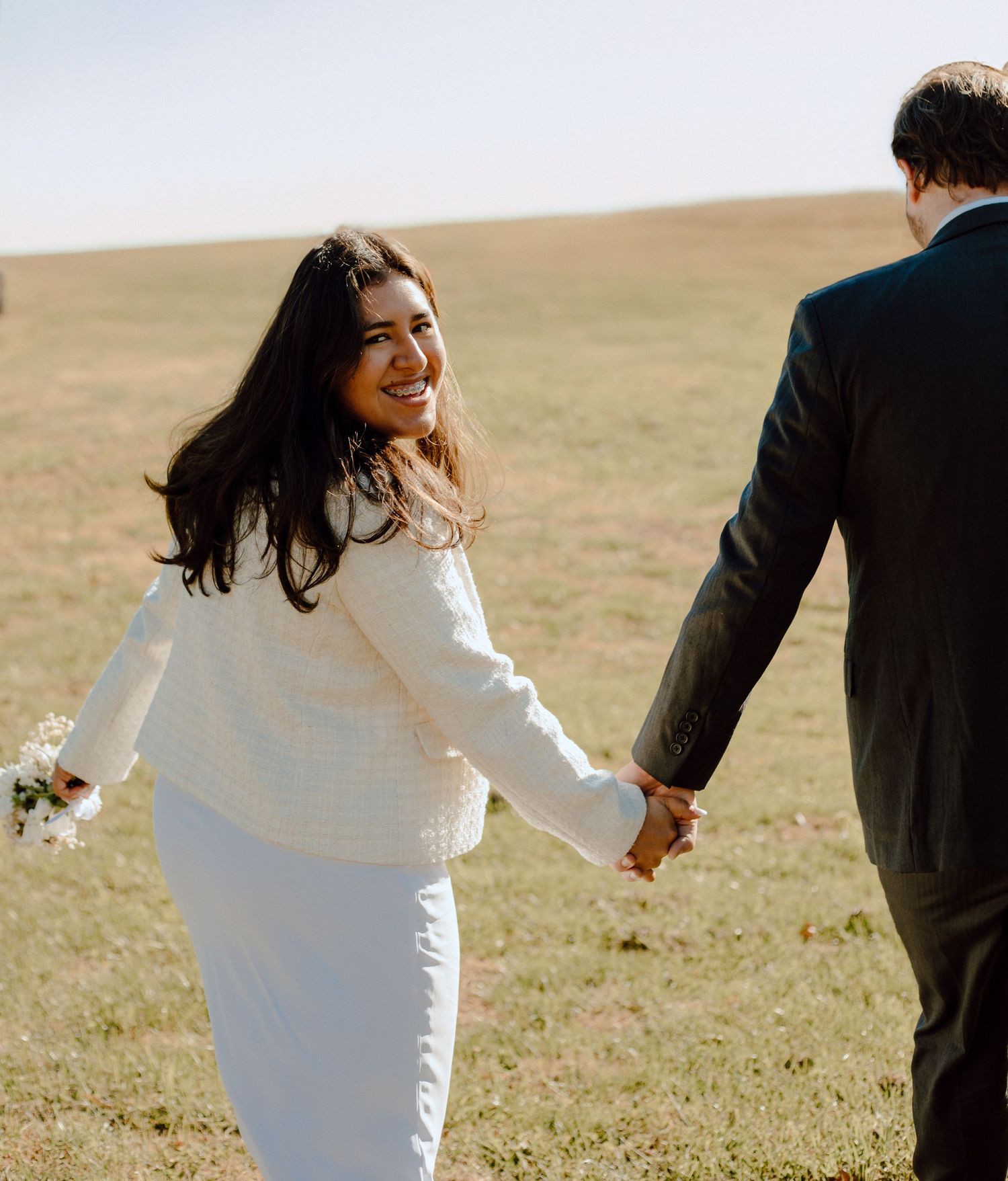 Couple in wedding attire holding hands while walking through a golden field on a sunny day.