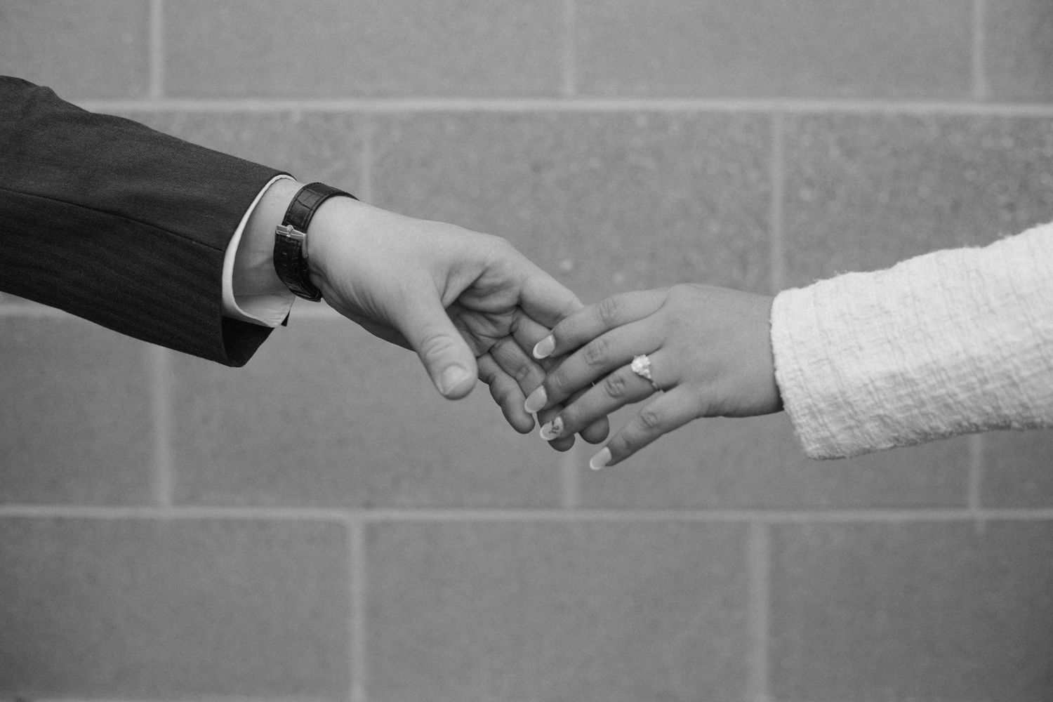 Two hands reaching towards each other in a gentle touch against a gray wall background, captured in black and white.