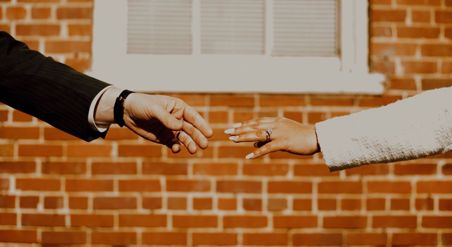 Two hands in formal attire reaching towards each other against a brick wall backdrop.