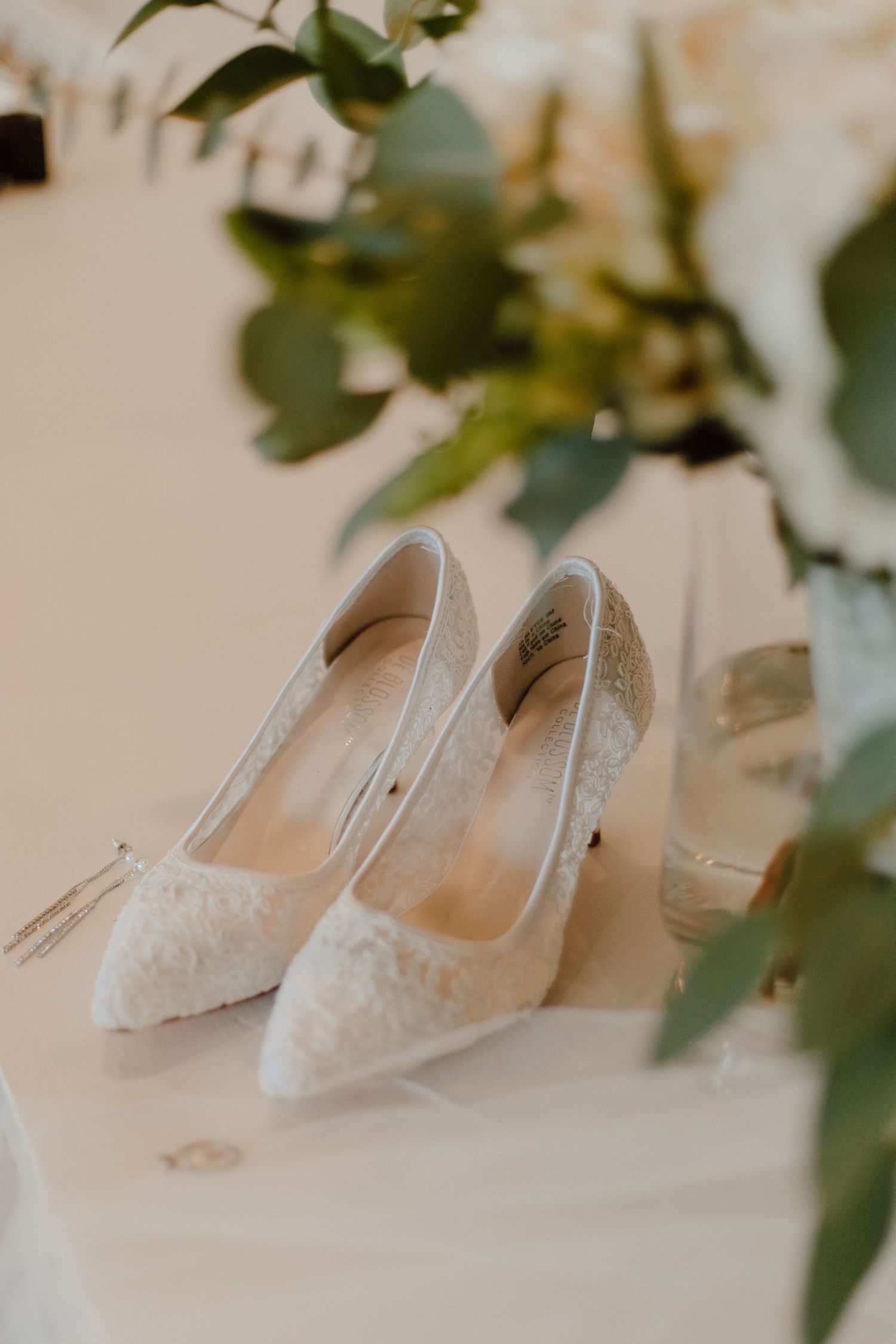 Elegant white lace wedding heels displayed on a table with green foliage in soft focus.