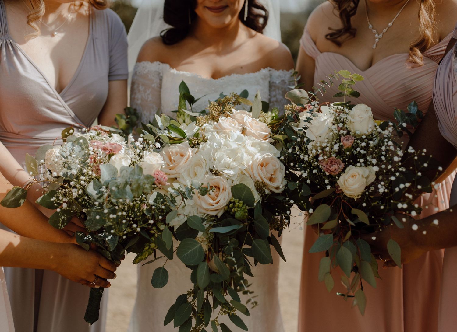 Elegant wedding bouquets with white roses, eucalyptus leaves and baby's breath held against blush pink dresses.