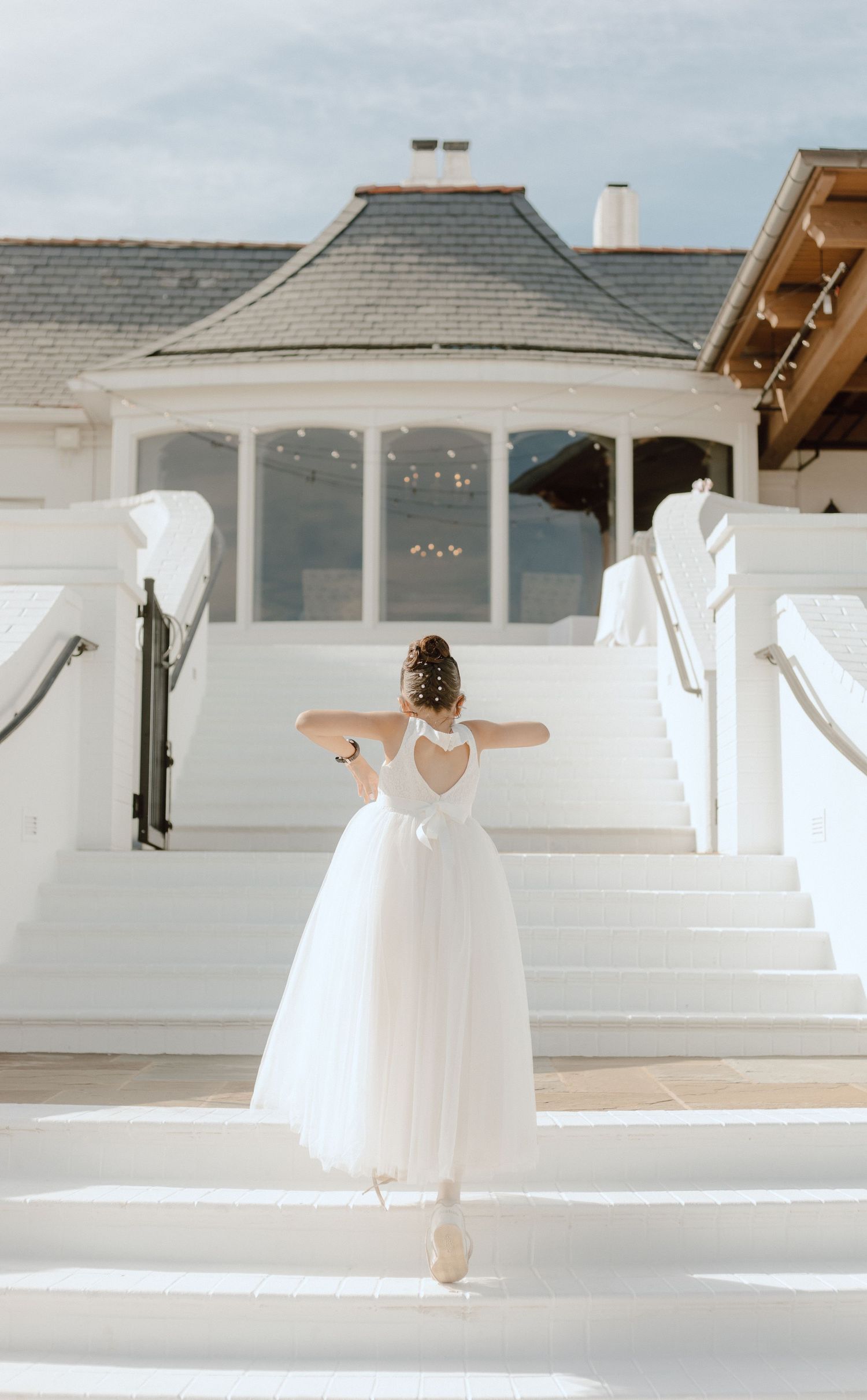 A ballet dancer in a white tulle dress poses gracefully on white steps in front of a gazebo-style building.