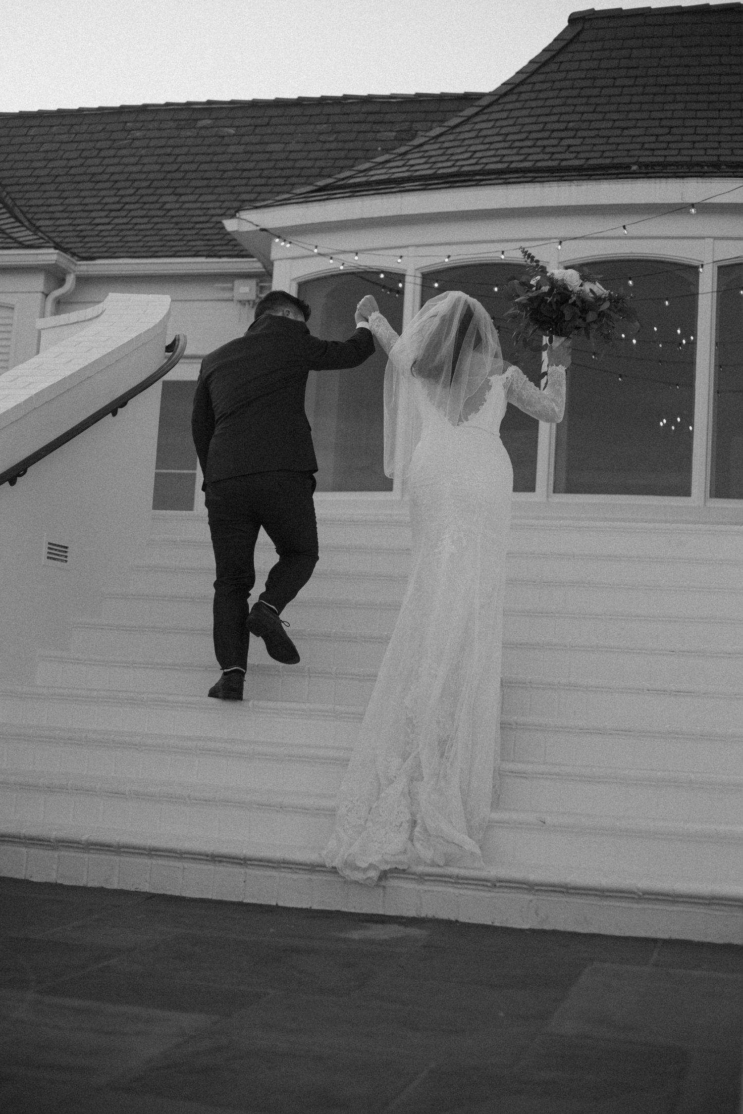 A couple ascends white porch steps in wedding attire during a candid black and white moment.