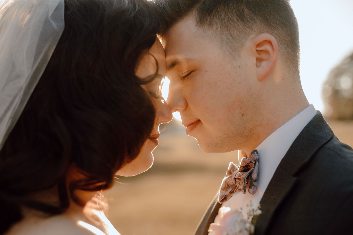 A tender moment between newlyweds touching foreheads in warm sunlight, with the bride wearing a veil.