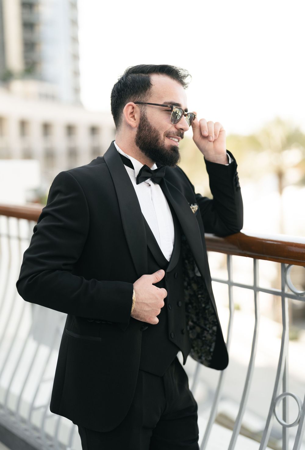 Stylish groom in black tuxedo and bowtie poses on outdoor balcony.