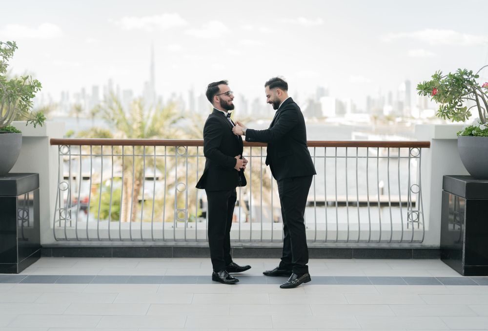 Two men in black suits stand face to face on a balcony with a cityscape in the background.