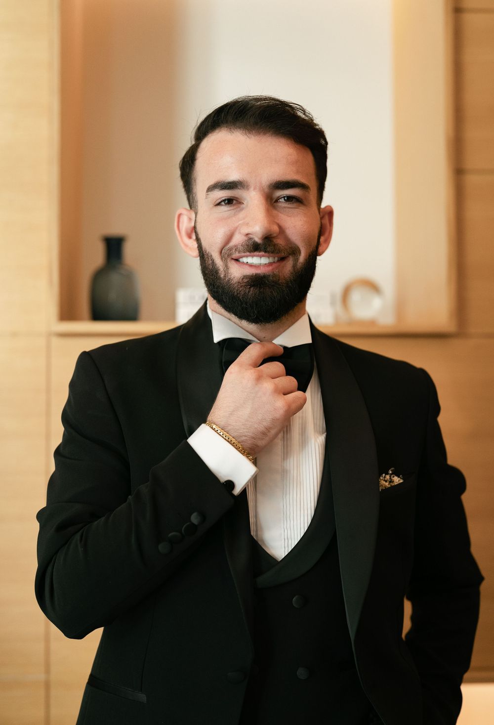 A groom adjusts his bowtie while wearing a classic black tuxedo with vest in an elegant indoor setting.