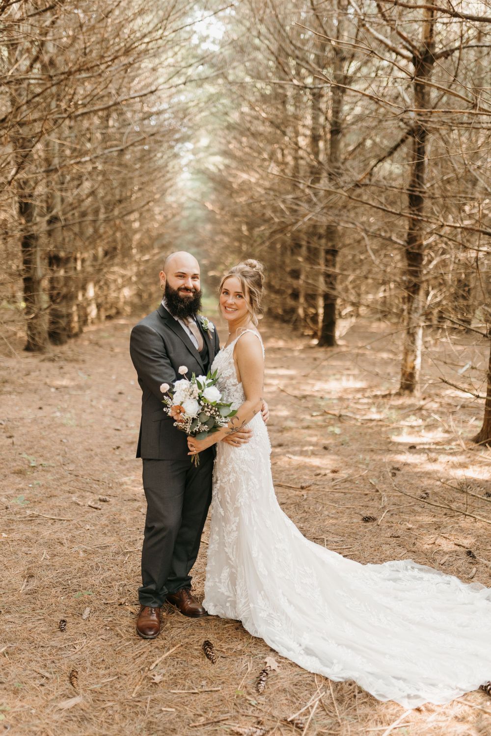 Bride in lace gown and bearded groom pose on pine needle path in wooded forest.