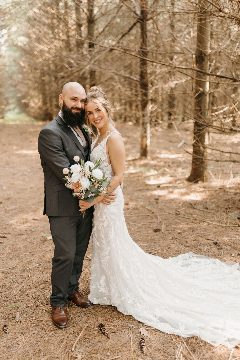 Bride holding bouquet and bearded groom embrace warmly in sunlit pine forest setting.
