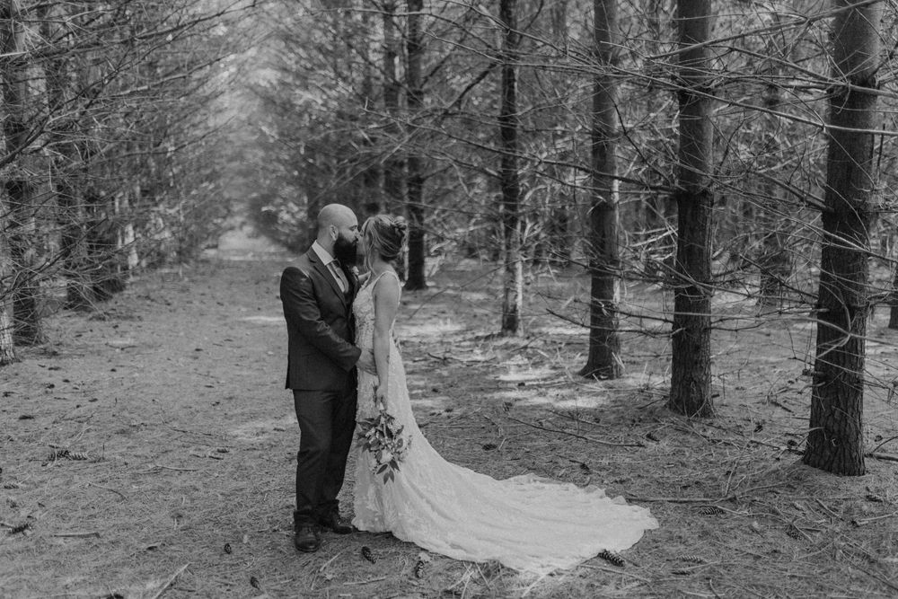 Black and white photo of bride and groom kissing on a forest path, bride wearing lace gown with long train.