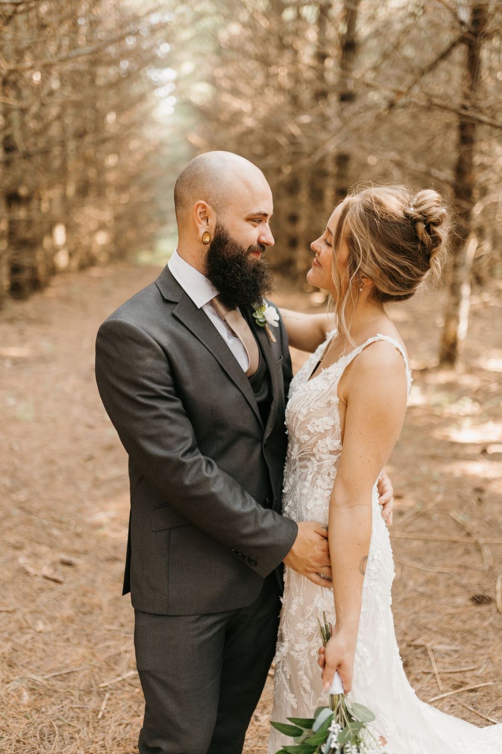 Groom in dark suit gazes lovingly at smiling bride in lace wedding dress among tall trees in forest.