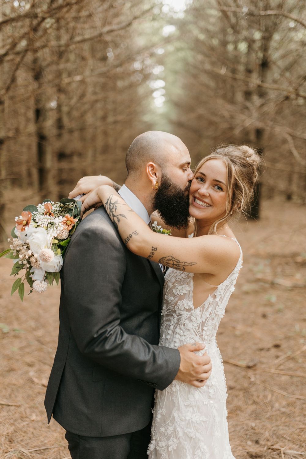 Tattooed groom kisses bride on cheek as she smiles holding a floral bouquet in a wooded forest setting.