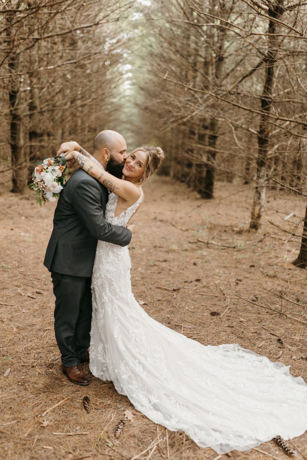 Groom embraces bride from behind in forest aisle, bride smiling holding bouquet with lace gown train flowing.