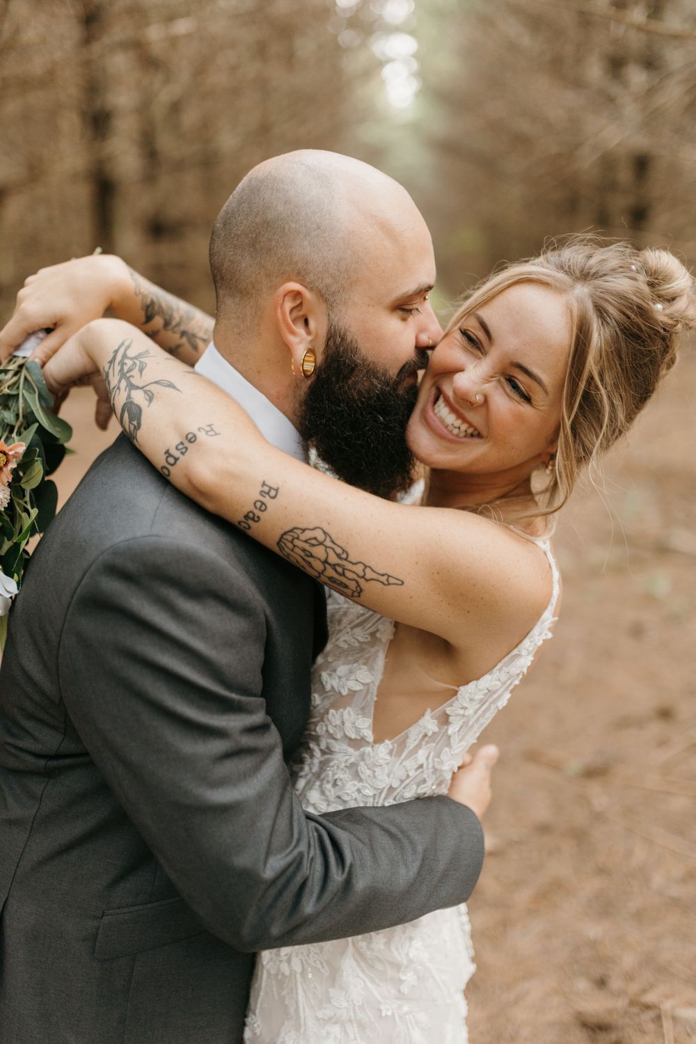 Laughing bride hugs tattooed groom around neck as he kisses her cheek during outdoor forest wedding portraits.