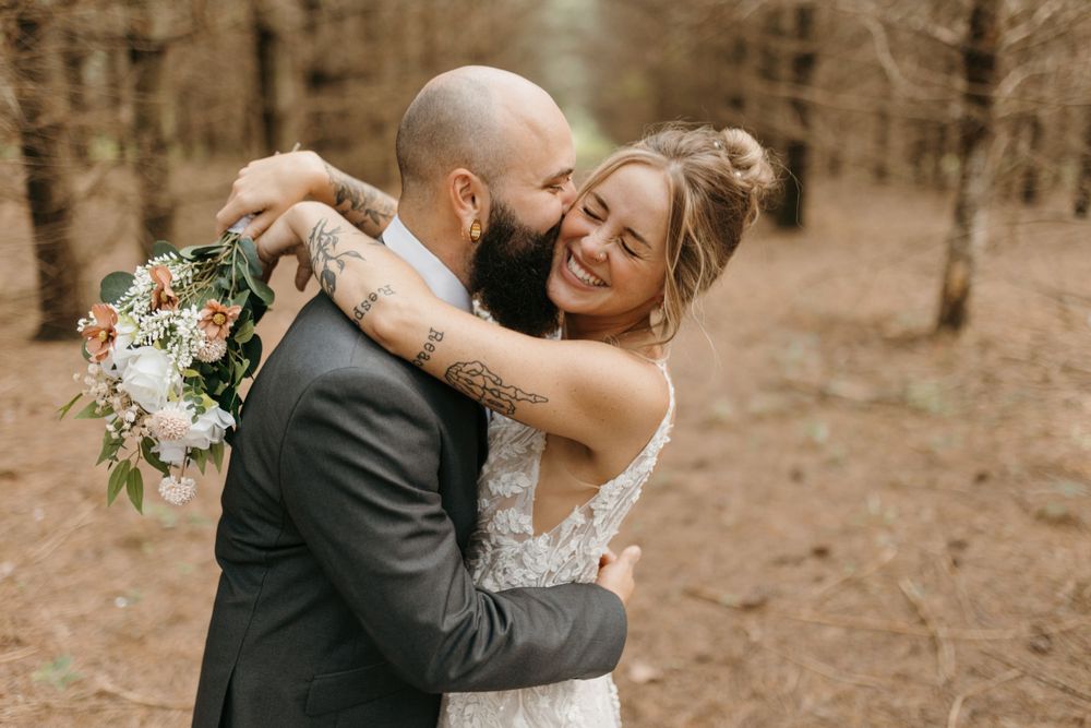 Bride and groom share a joyful embrace in a sunlit forest, bride holding a pastel floral bouquet.