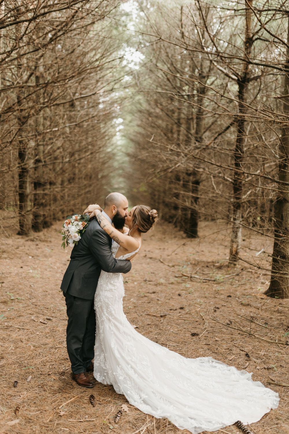 Bride and groom share a romantic kiss while embracing on pine needle covered forest floor, lace train spread out.