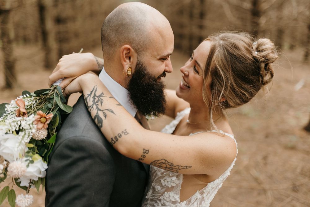 Bride and groom laugh together face to face in forest, bride holding white and peach floral wedding bouquet.