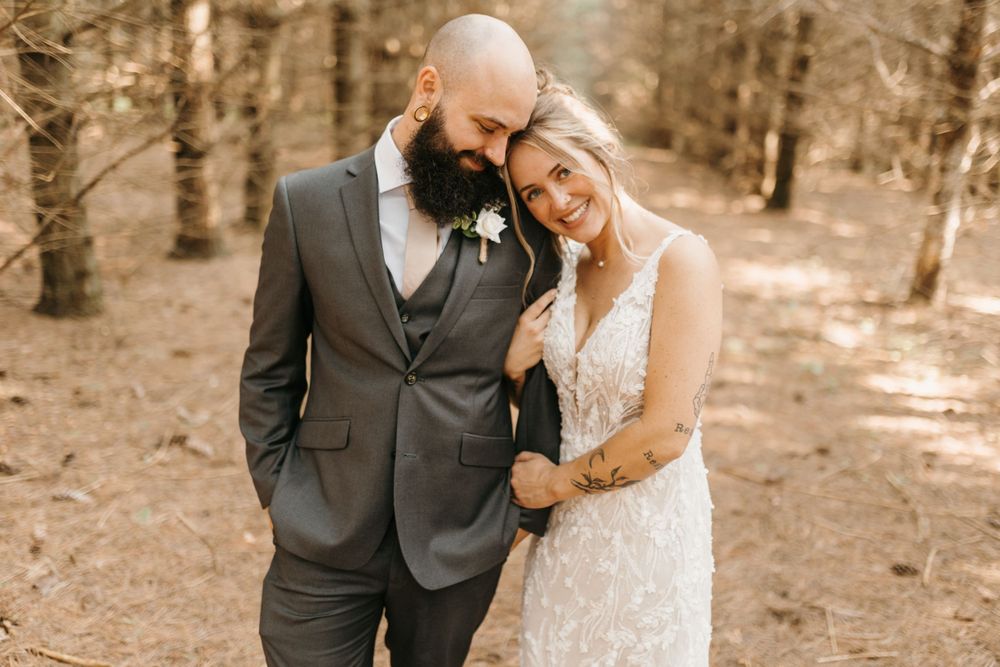 Groom leans head toward smiling bride as she holds his arm in a warm sunlit forest setting.
