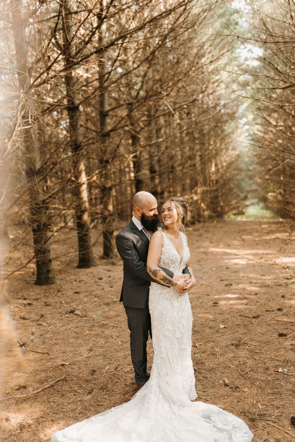 Bride and groom embrace on a pine forest path, bride in lace gown with train, groom in dark suit.