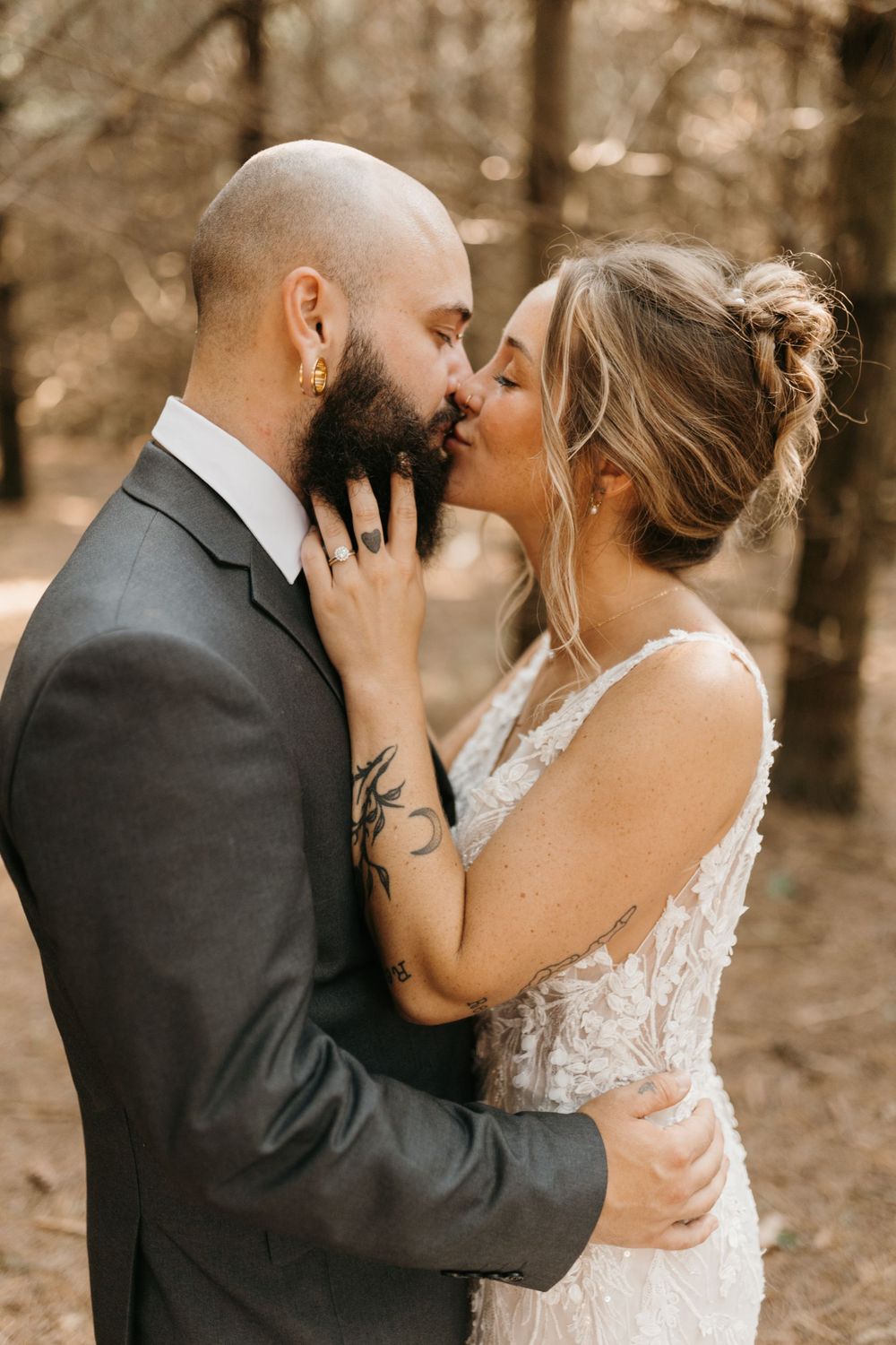 Bride and groom share a romantic kiss outdoors, groom with beard and tattoos, bride in lace wedding dress.