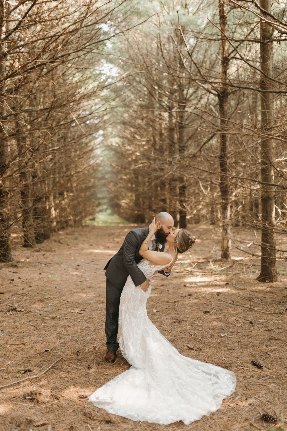 Groom dips bride for a kiss on a pine tree-lined forest path, bride in flowing lace gown with train.