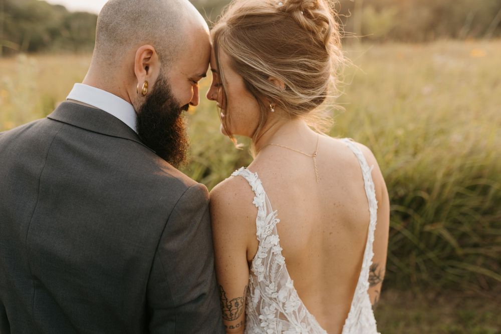 Bride and groom touch foreheads tenderly in golden field, bride in low-back lace gown with tattoo visible.