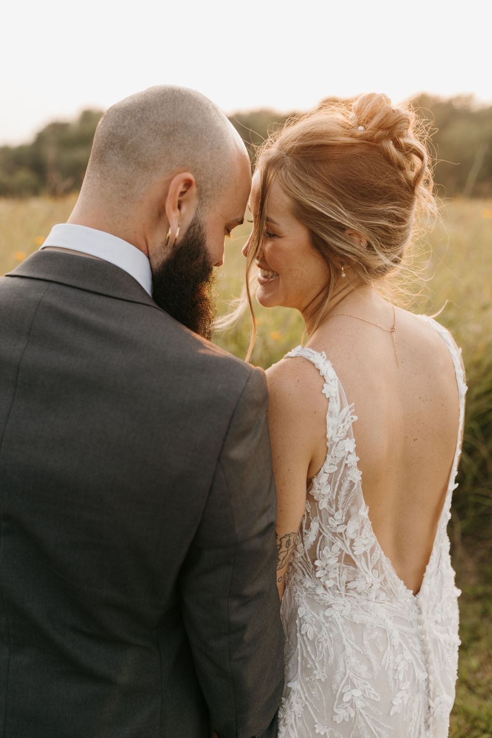 Smiling bride and bearded groom share an intimate moment in a sunlit meadow, bride in lace backless gown.