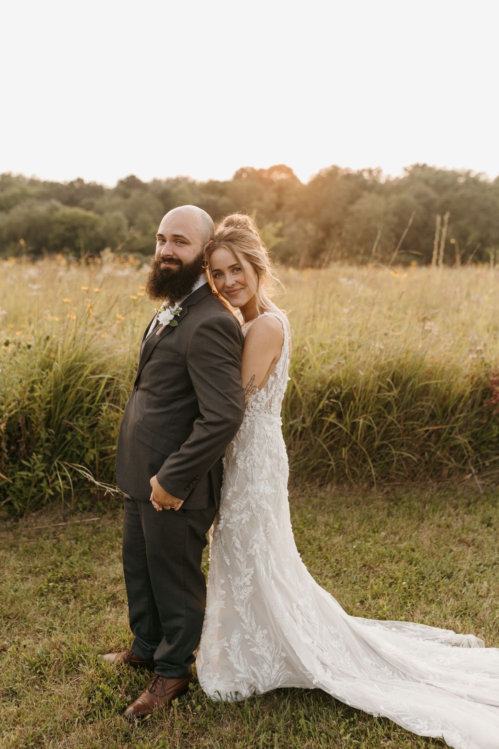Bride and groom pose back-to-back in golden hour meadow, bride in lace gown with long train, both smiling.