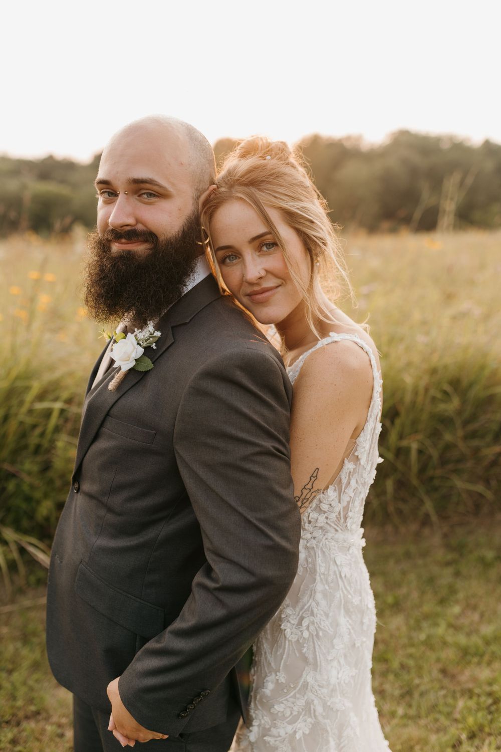 Bride leans on groom's shoulder in sunset meadow, both smiling at camera, bride in floral lace wedding gown.