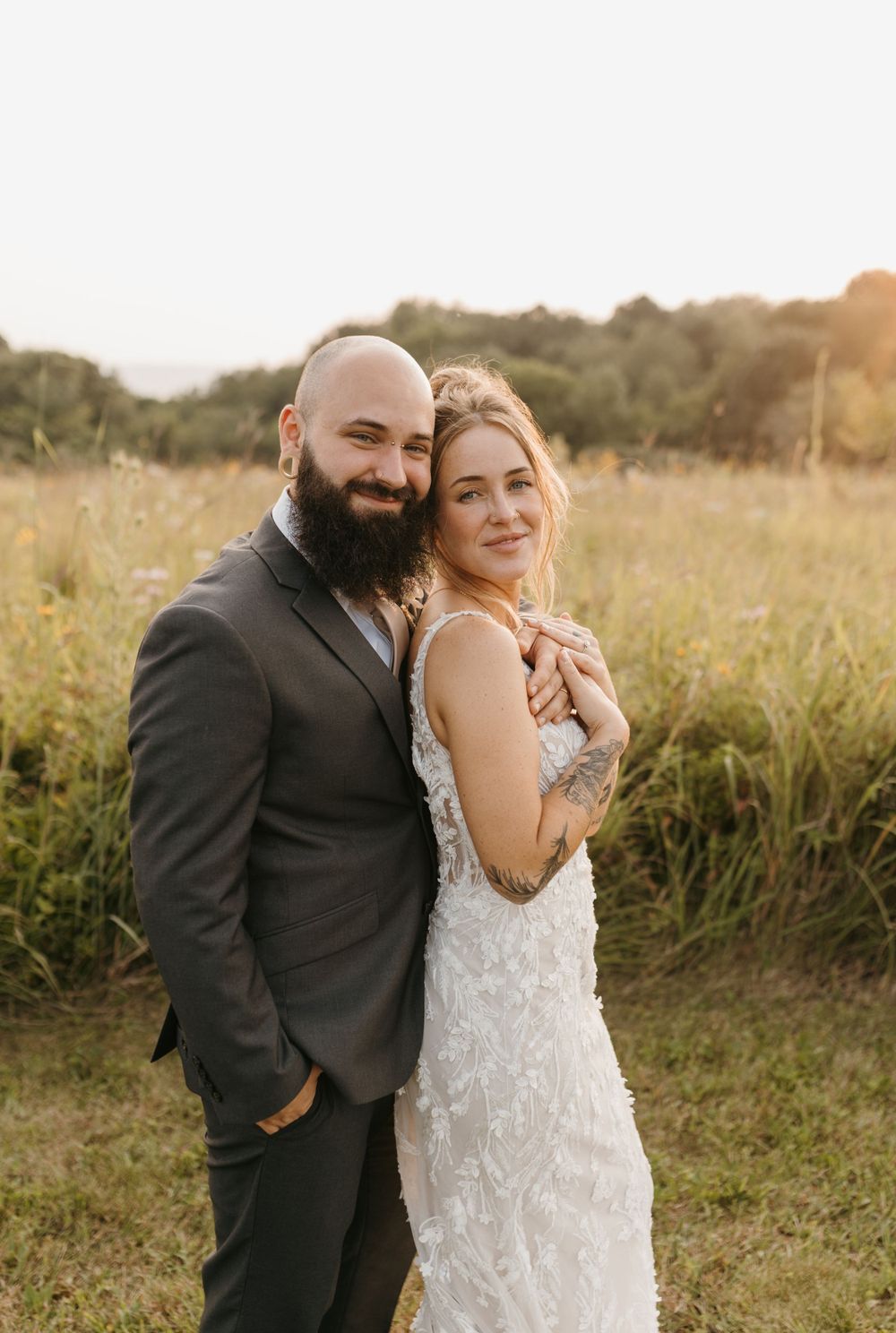 Bride embraces groom from behind in golden field, both smiling at camera, bride in lace applique wedding dress.