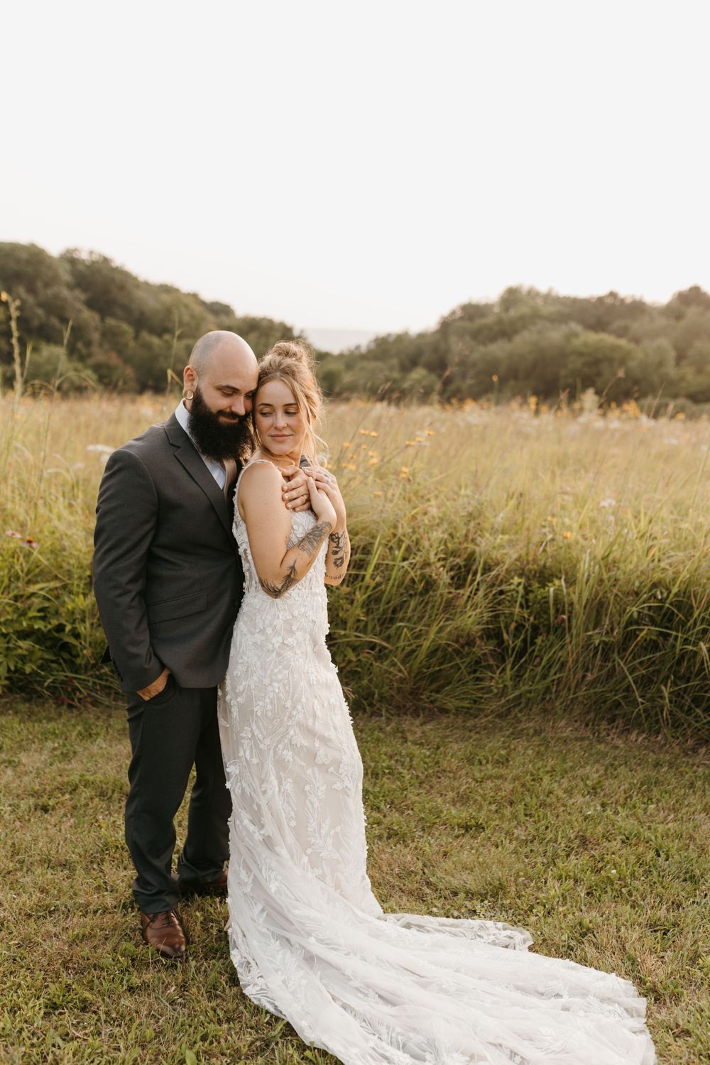 Groom embraces bride from behind in sunlit meadow, bride in lace gown with long train, both eyes closed.