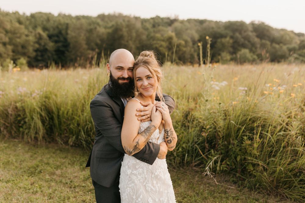 Bearded groom hugs bride playfully in golden meadow, bride laughing in lace wedding dress at sunset.