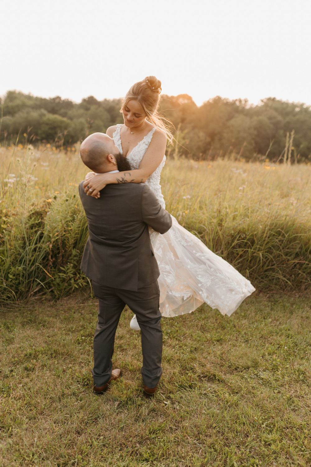 Groom lifting bride in white lace dress during golden hour in a meadow.