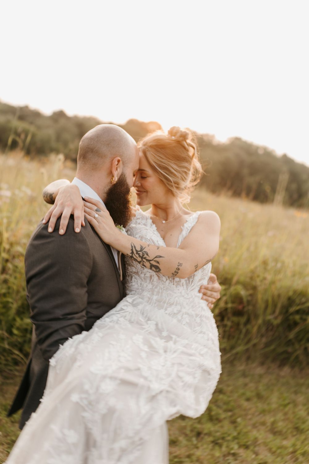 Groom carrying bride in lace wedding gown at sunset in a field.