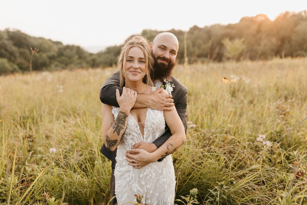Groom embracing bride from behind in a wildflower meadow at sunset.