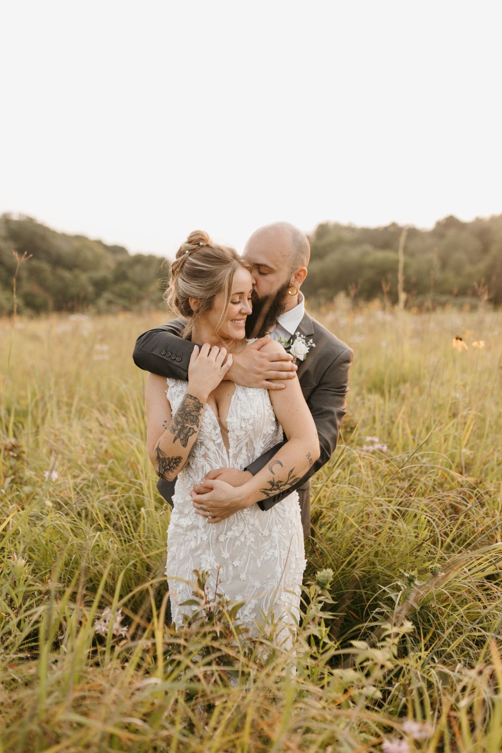 Groom tenderly embracing bride from behind in tall golden grass.