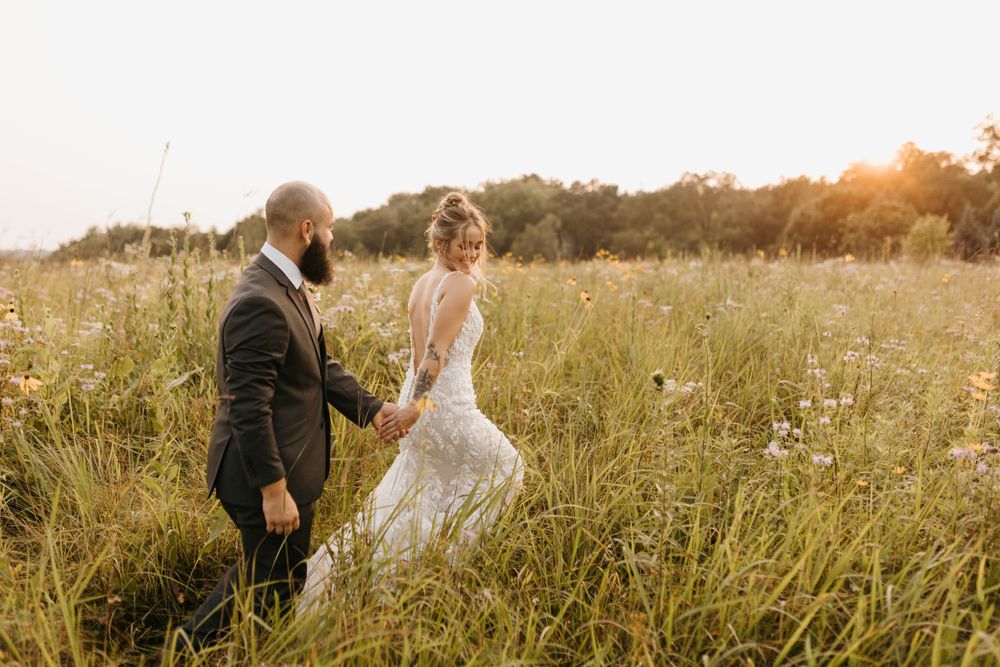 Bride and groom walking hand in hand through a sunlit meadow.