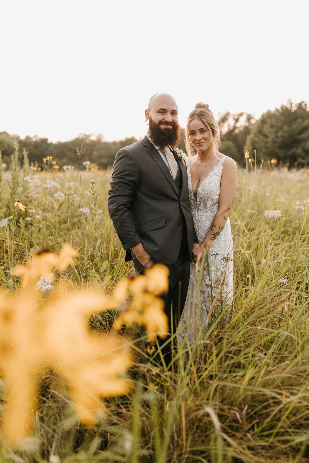 Bride and groom smiling in a wildflower meadow, she in lace gown, he in dark suit, golden hour light.