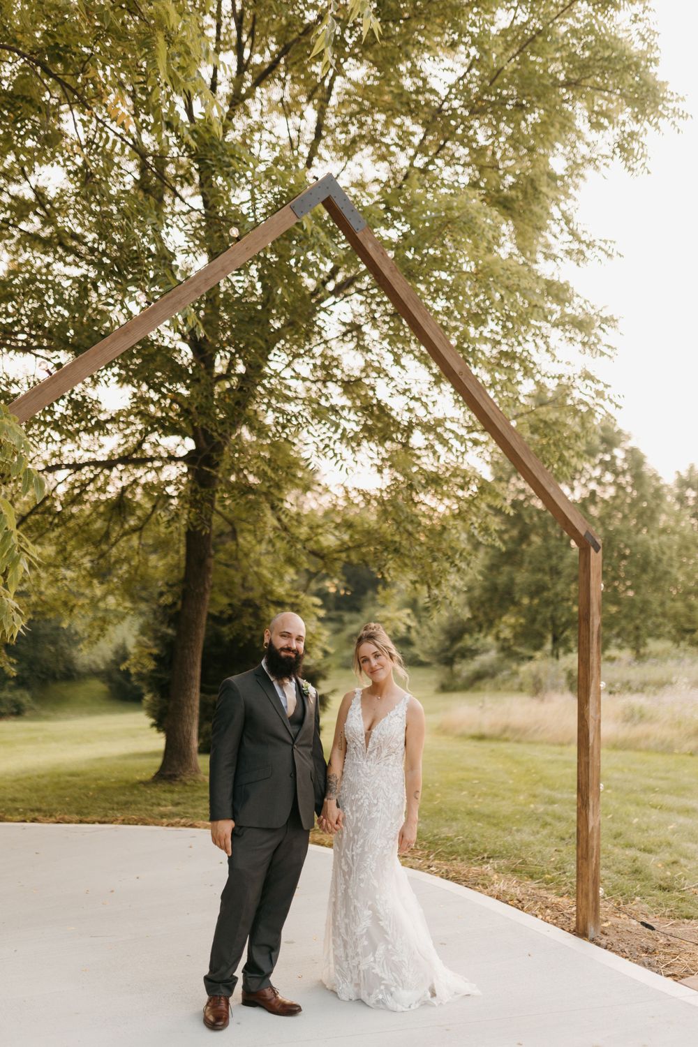 Bride and groom posing under a modern wooden arch ceremony structure surrounded by lush green trees.