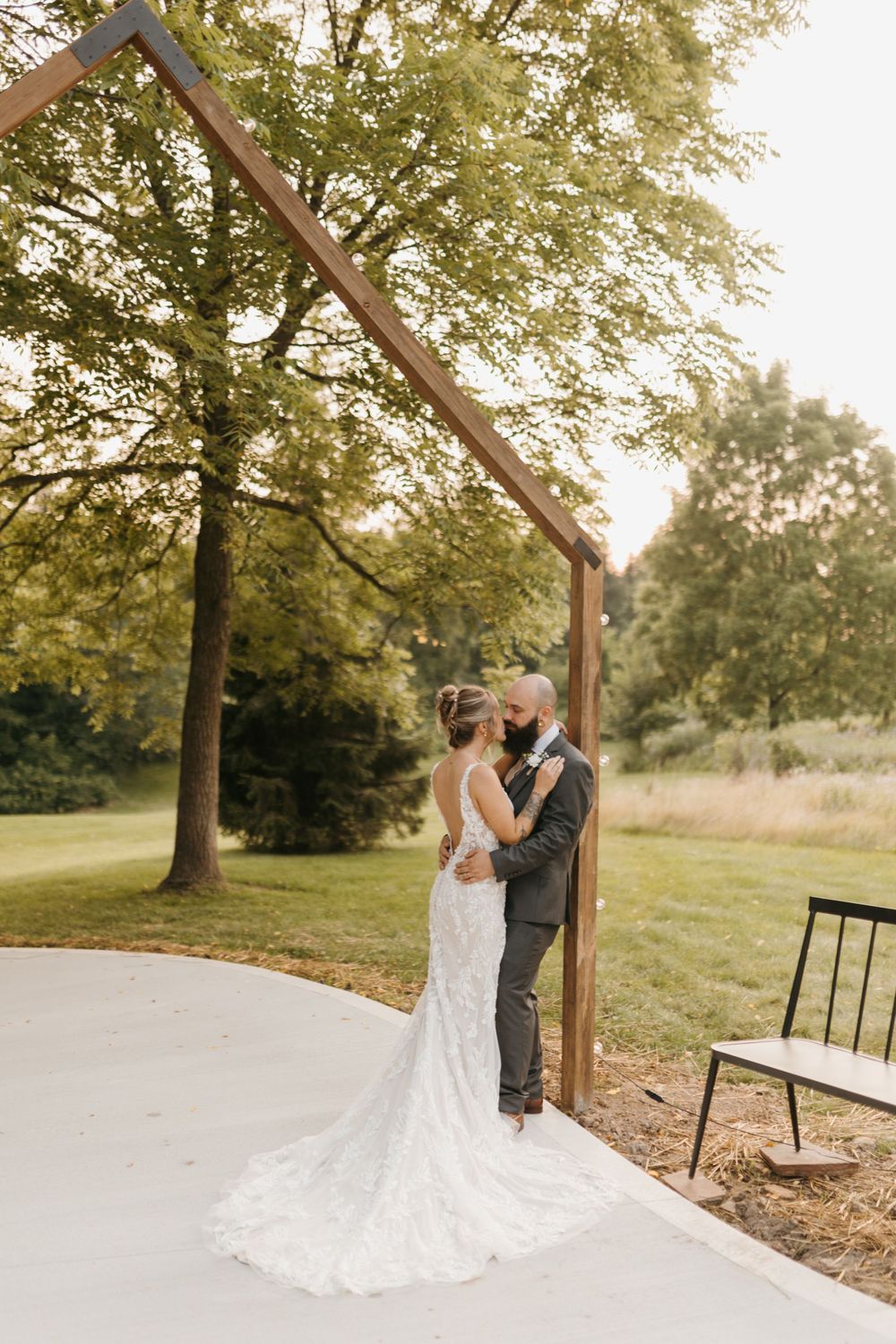 Bride in lace gown and groom embracing under a minimalist wooden arch at an outdoor wedding venue.