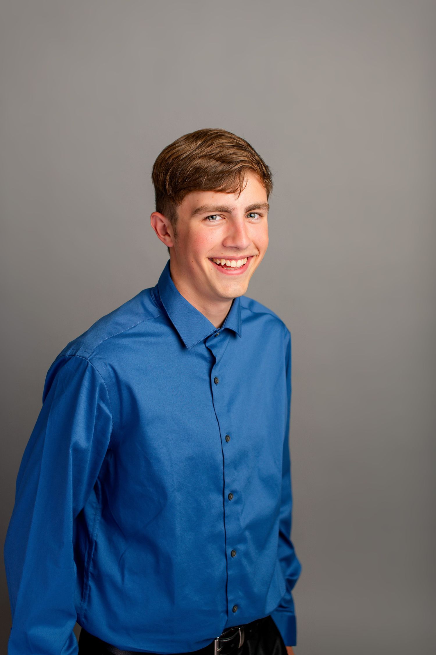 A smiling young man with brown hair wearing a blue collared shirt stands against a gray background.